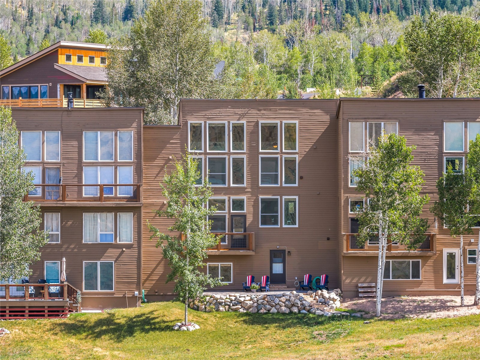 Brown multi-story condominium with balconies, outdoor chairs, and a forested mountain backdrop.