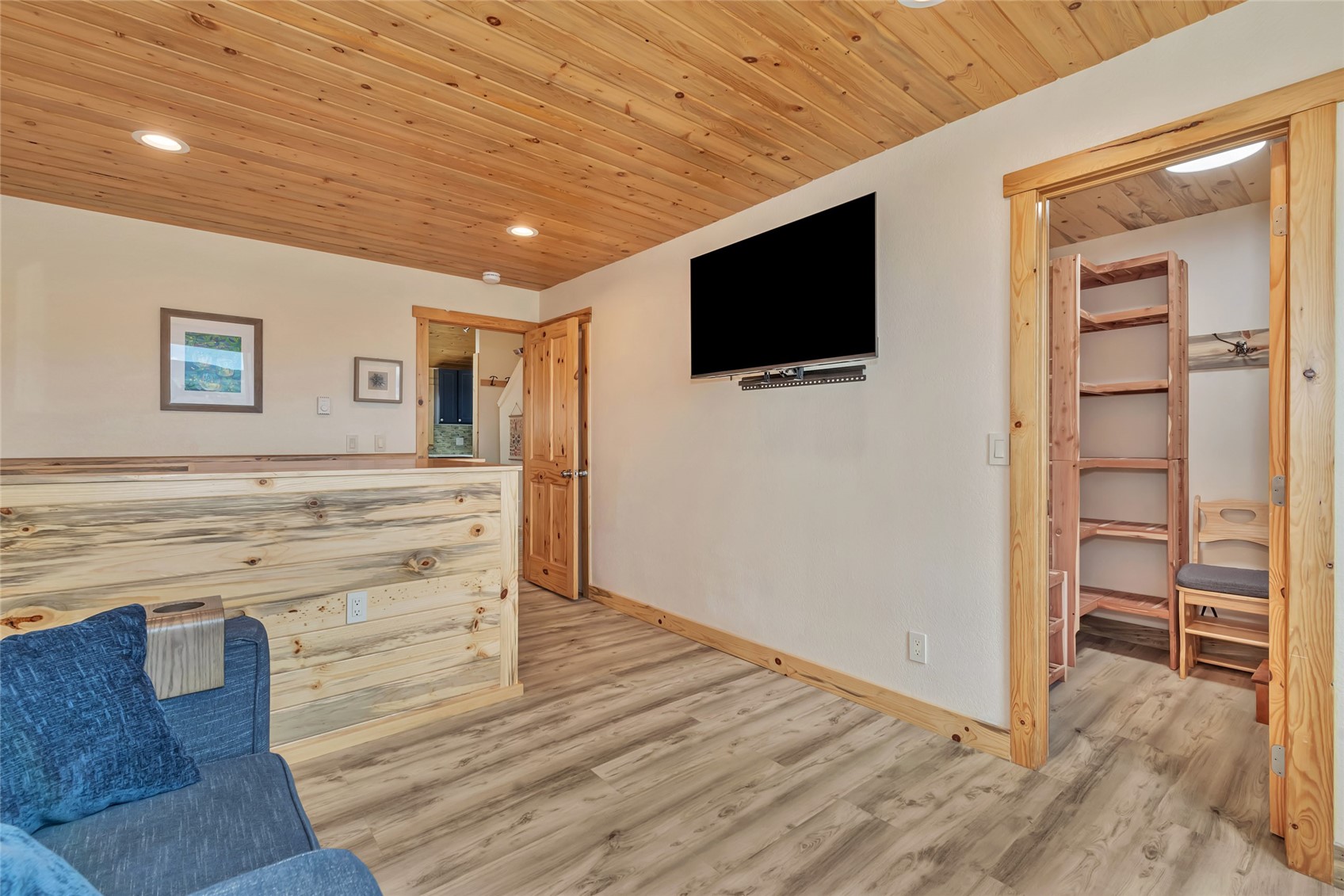 Furnished room with a blue sofa, wall-mounted TV, knotty pine ceiling, and open doorway to a room with shelving.