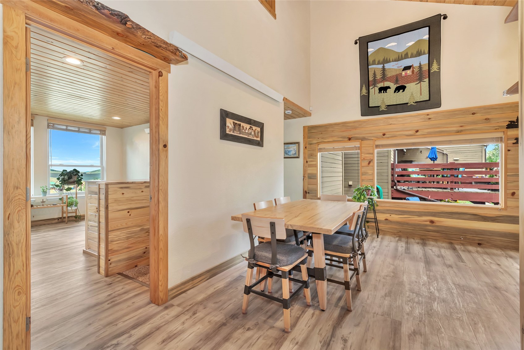 Dining area with a wooden table, knotty wood trim, and large windows overlooking a deck.