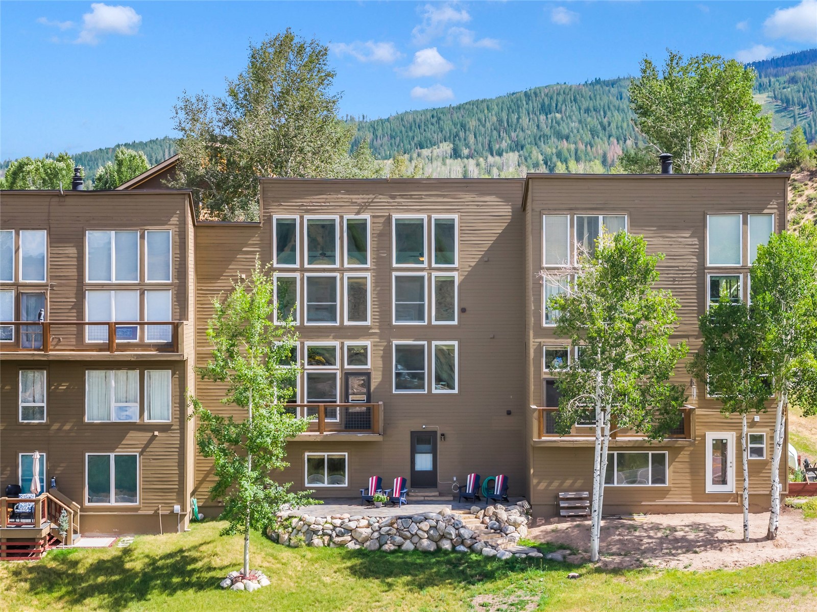 Brown multi-story condominium with balconies and a stone-edged patio, backed by a forested mountain.