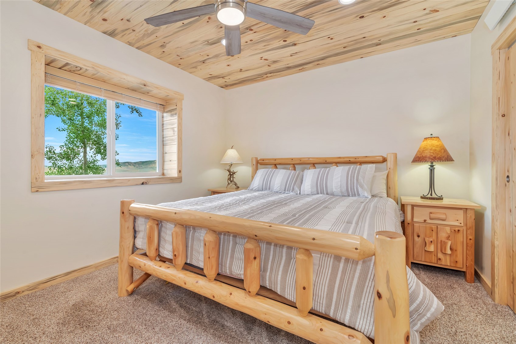 Bedroom with a rustic log bed, wood-paneled ceiling, and window overlooking trees and rolling hills.