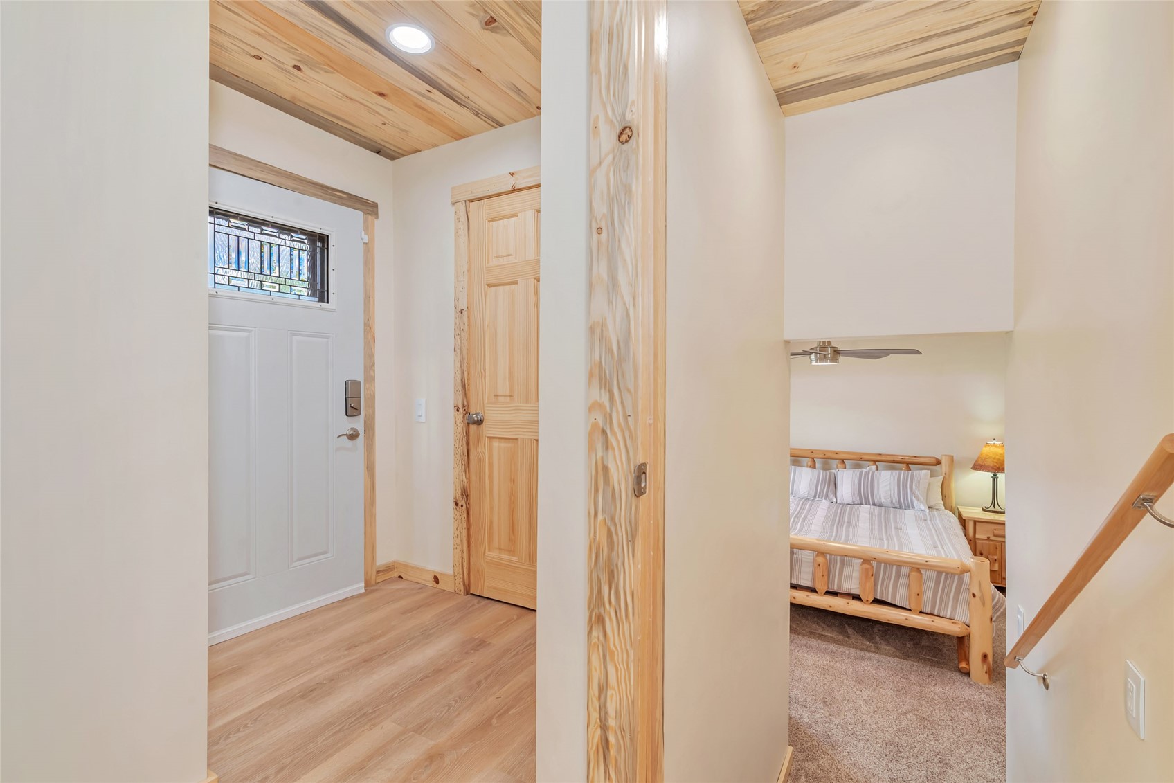 Hallway with wood-plank ceiling and glass-paneled entry door, looking into a bedroom with a wooden bed.