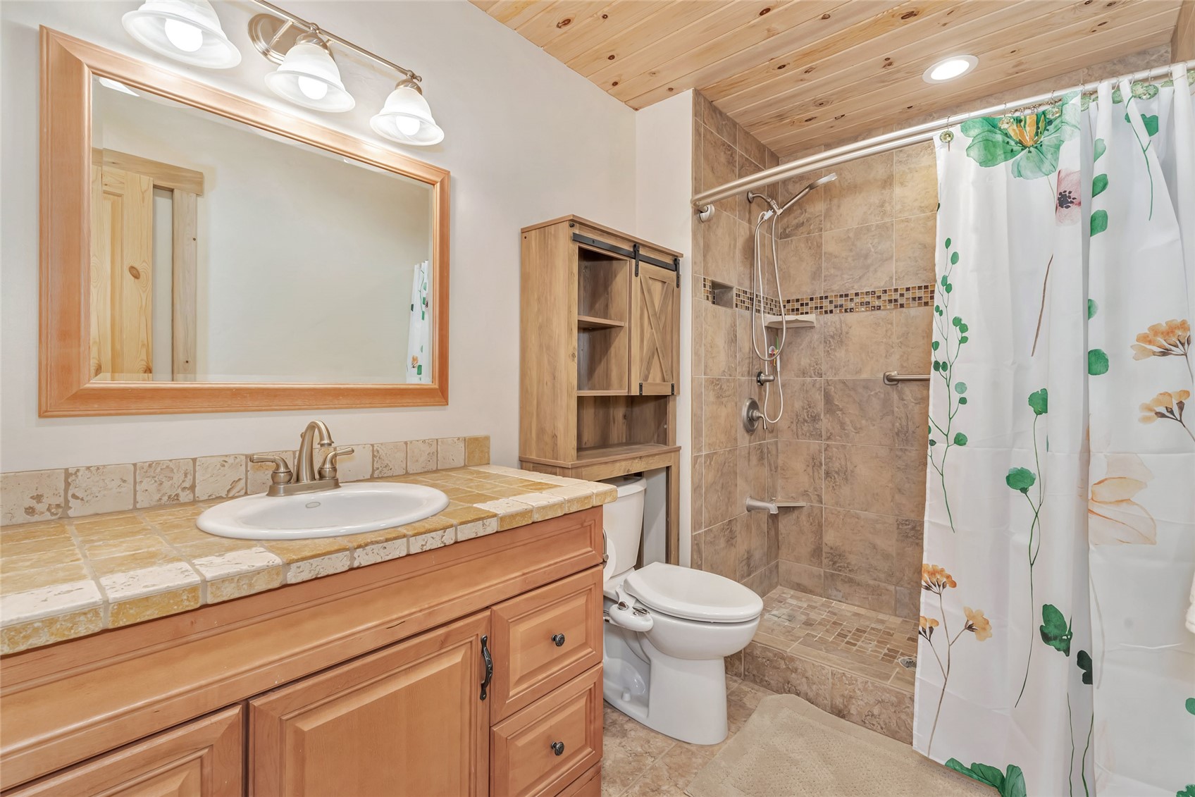 Bathroom with tiled shower, floral curtain, wood vanity, and plank ceiling.