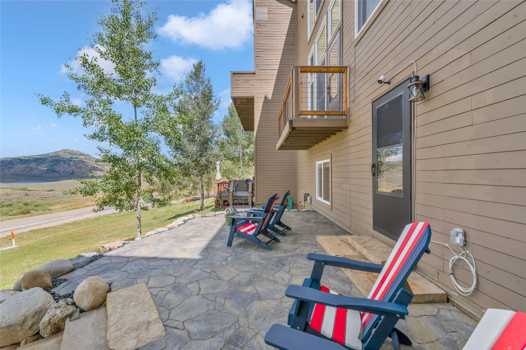 Stone patio with colorful Adirondack chairs beside a tan home, overlooking open land and a mountain ridge.