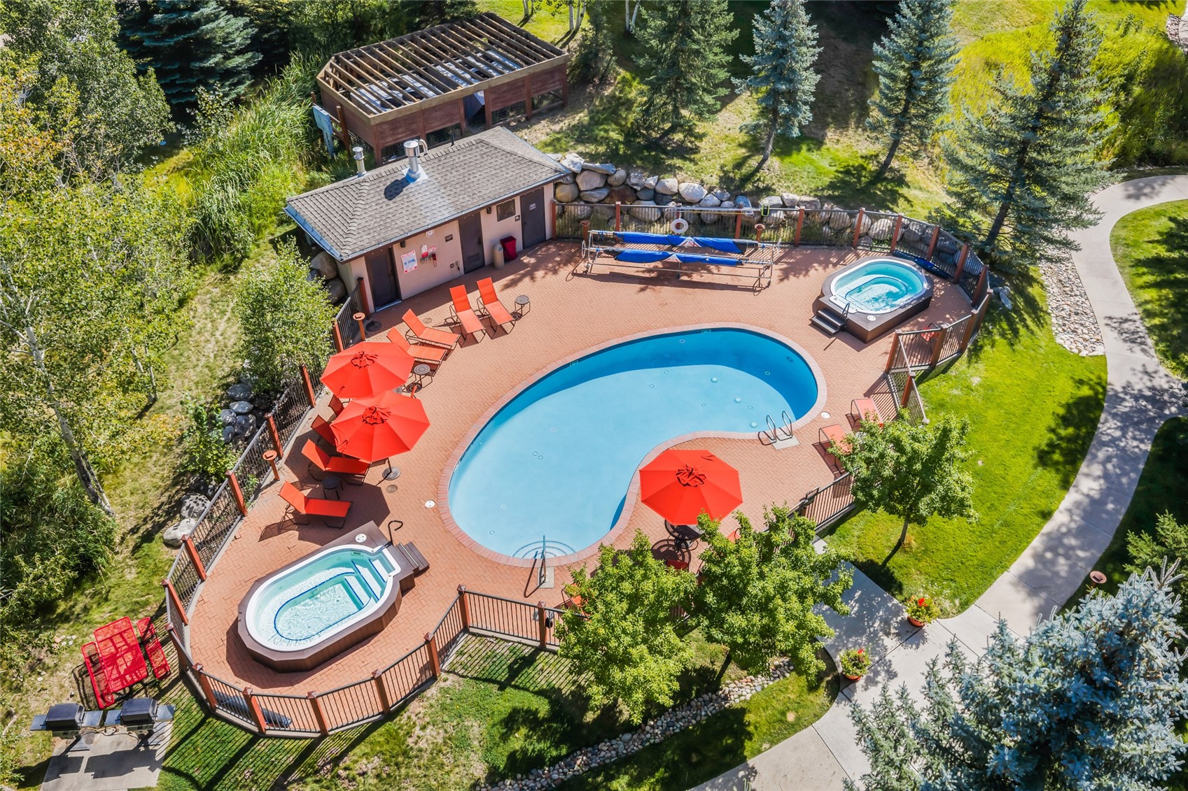 Outdoor pool and two hot tubs surrounded by a brick patio with red umbrellas and lounge chairs.