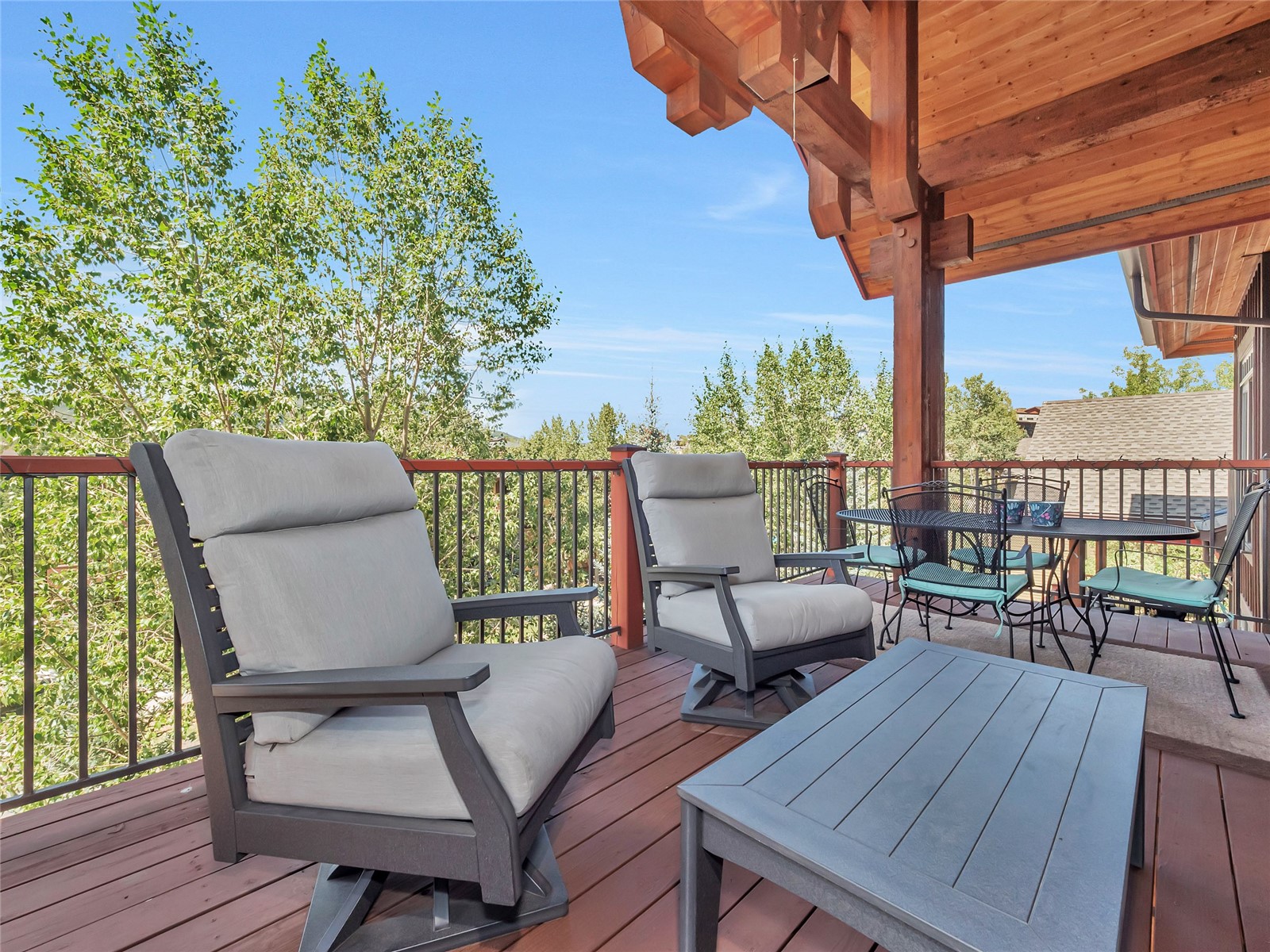 Furnished wooden deck with cushioned lounge chairs beneath a timber overhang, overlooking leafy trees.