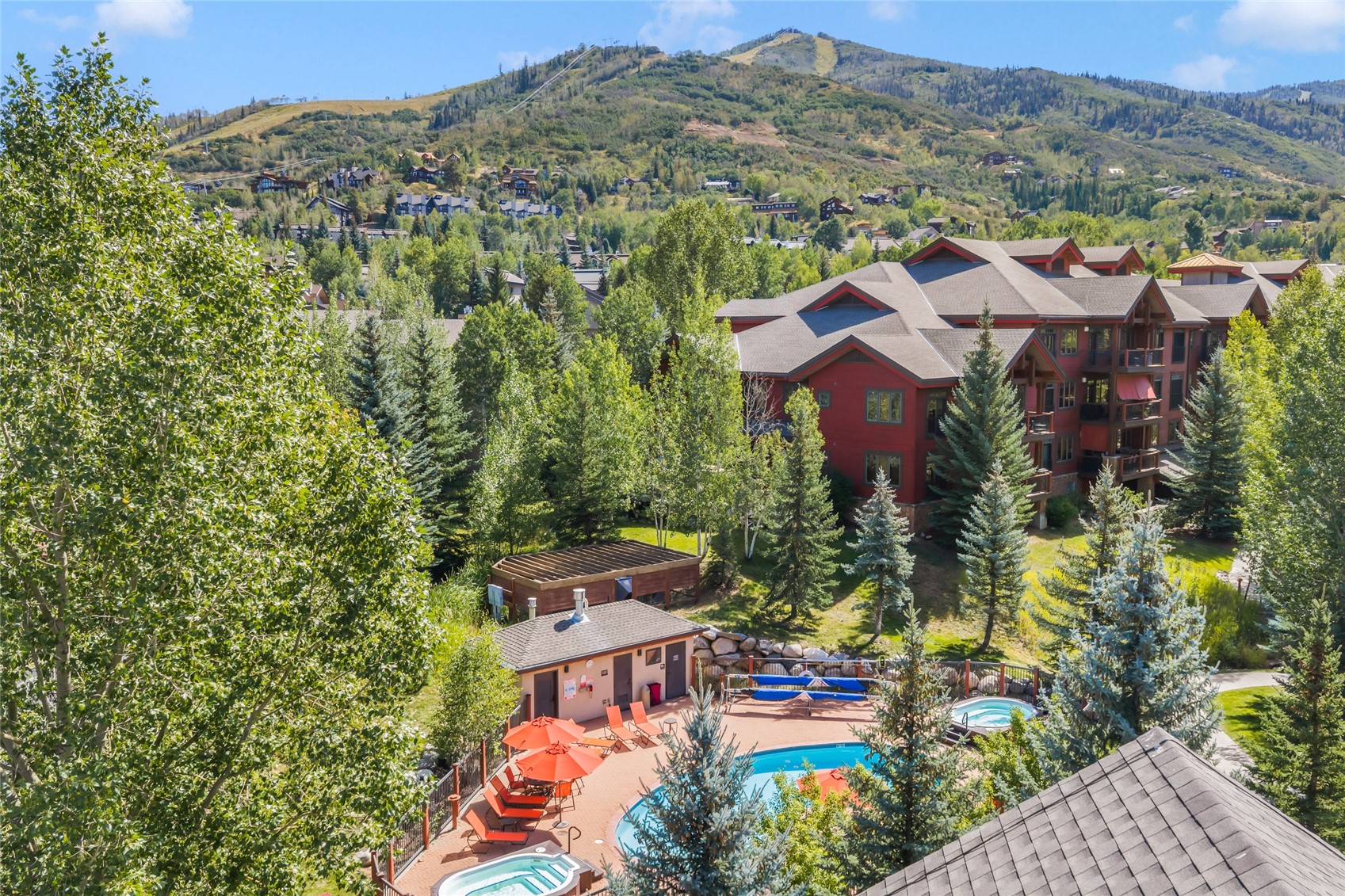 Aerial view of a mountain condominium complex with outdoor pools and a forested mountain backdrop.