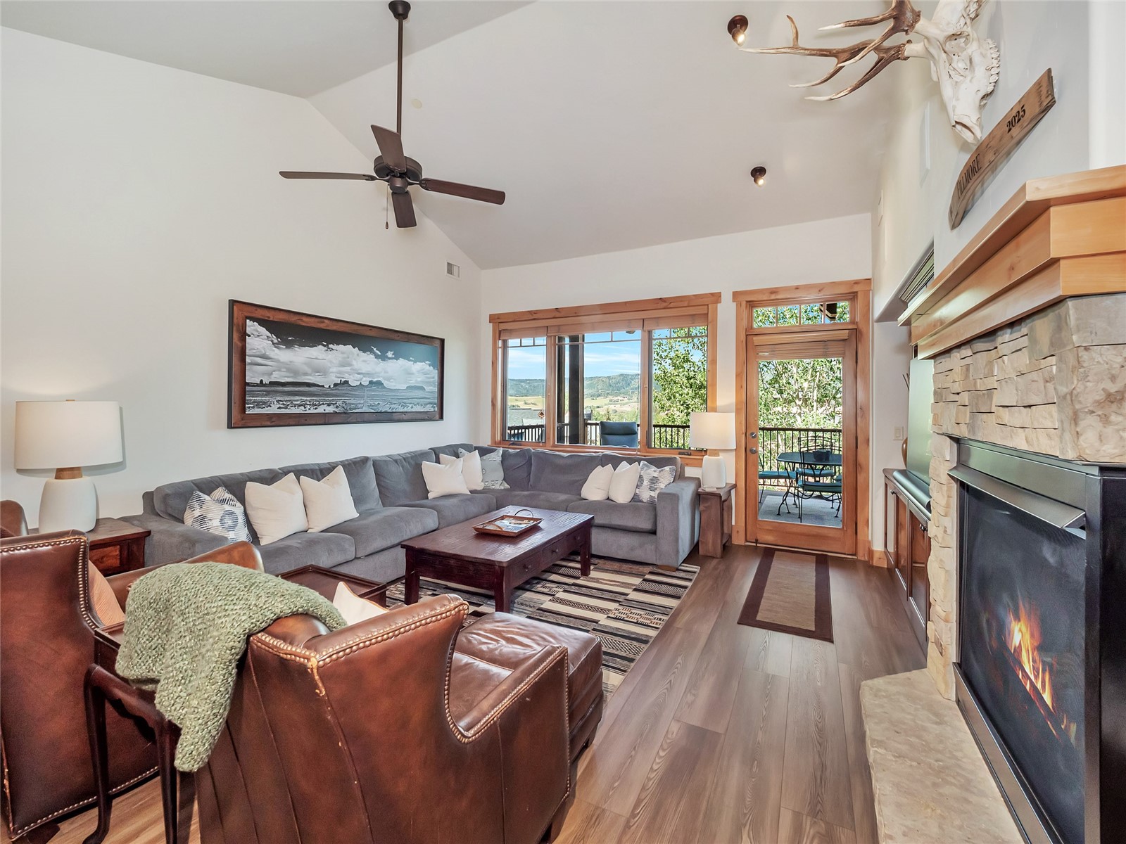 Living room with a stone fireplace, gray sectional, leather chairs, and balcony doors overlooking green hills.