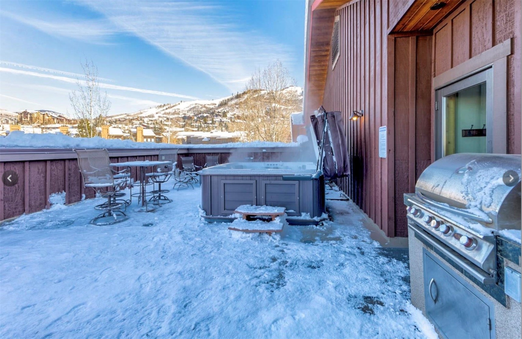 Snowy deck with a steaming hot tub, patio seating, and mountain views.