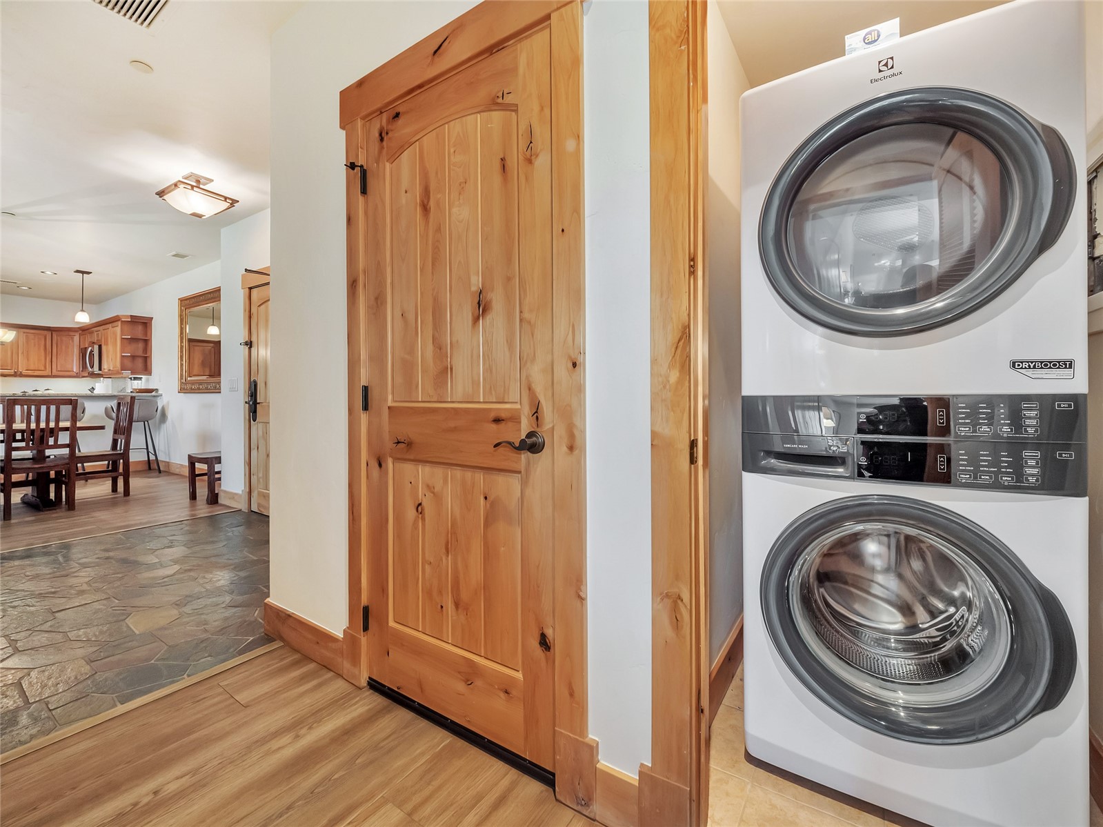 Laundry area with stacked washer and dryer beside a rustic wooden door; dining area visible beyond.