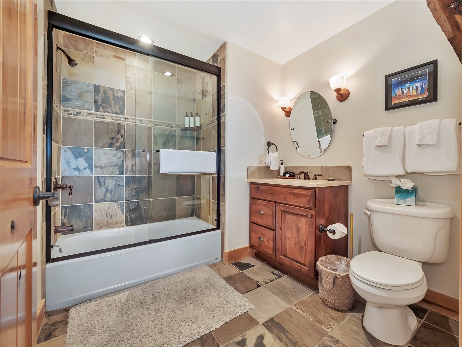 Bathroom with a glass-enclosed tiled tub, wood vanity, oval mirror, and toilet.