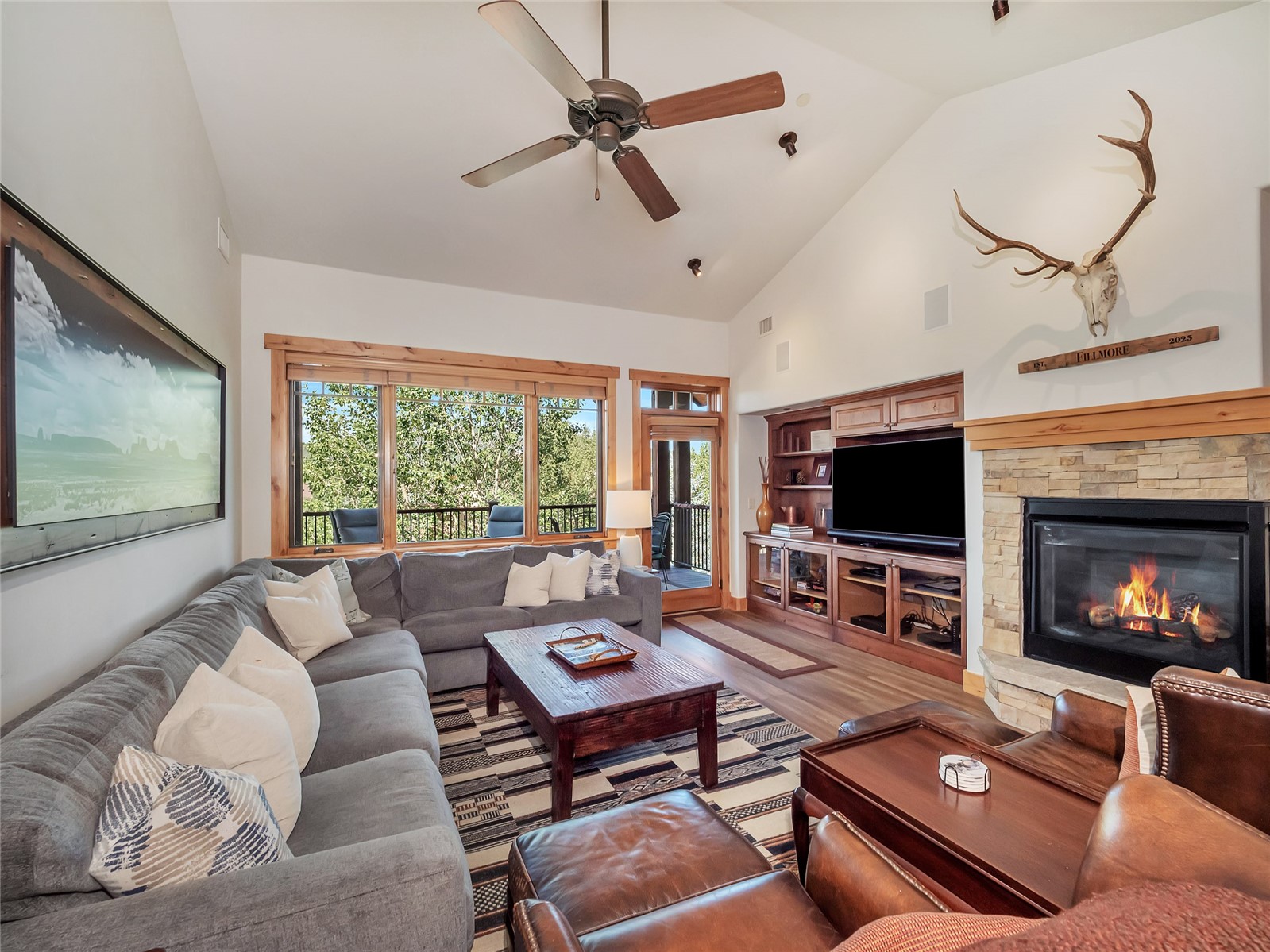 Living room with a gray sectional, stone fireplace, and large windows overlooking leafy trees.