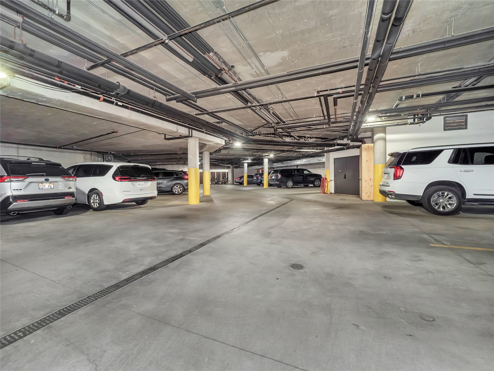 Underground parking garage with parked vehicles, yellow column guards, and exposed ceiling pipes.