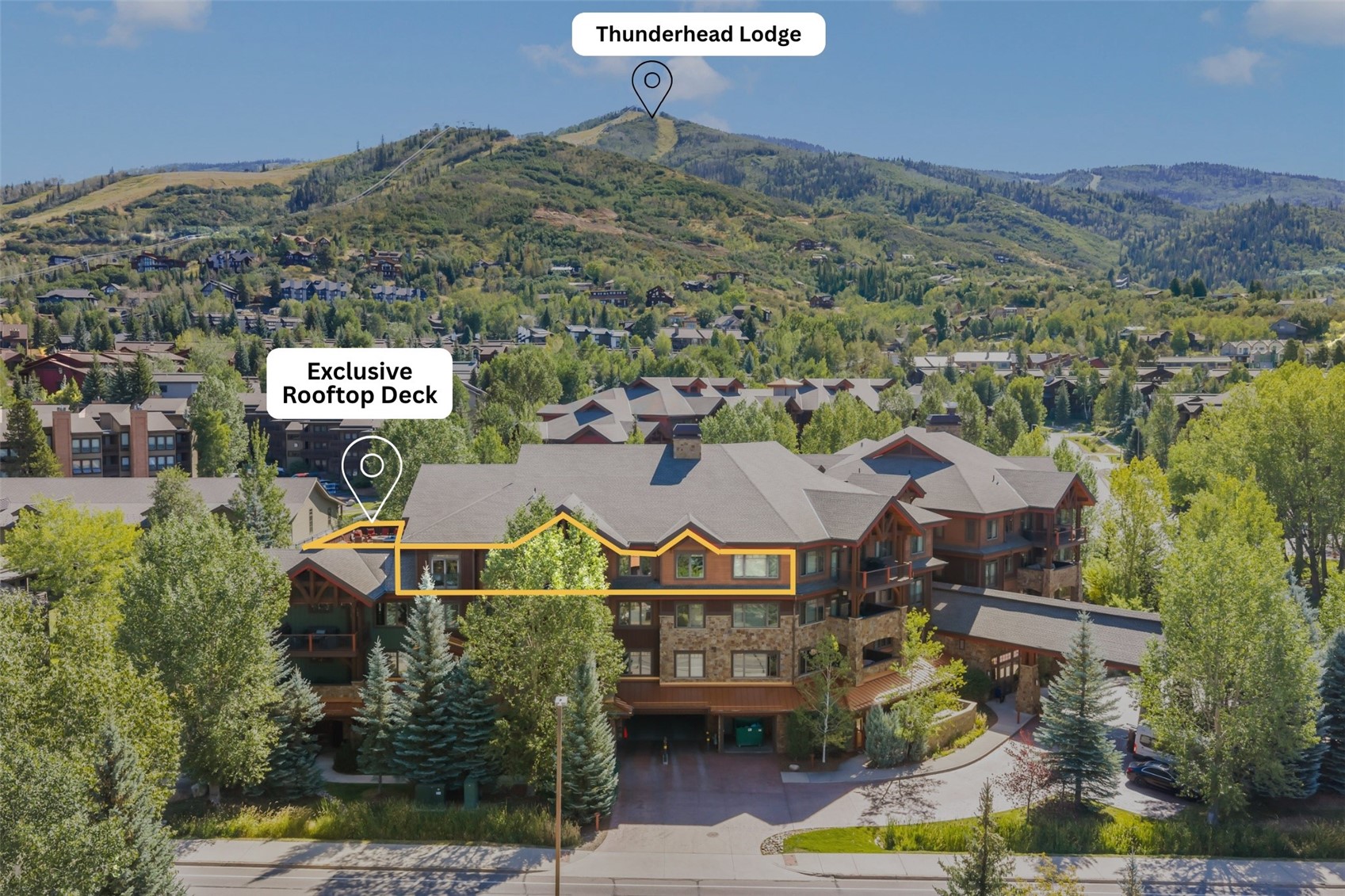 Lodge-style condominium building with an outlined rooftop deck, surrounded by trees and mountain scenery.
