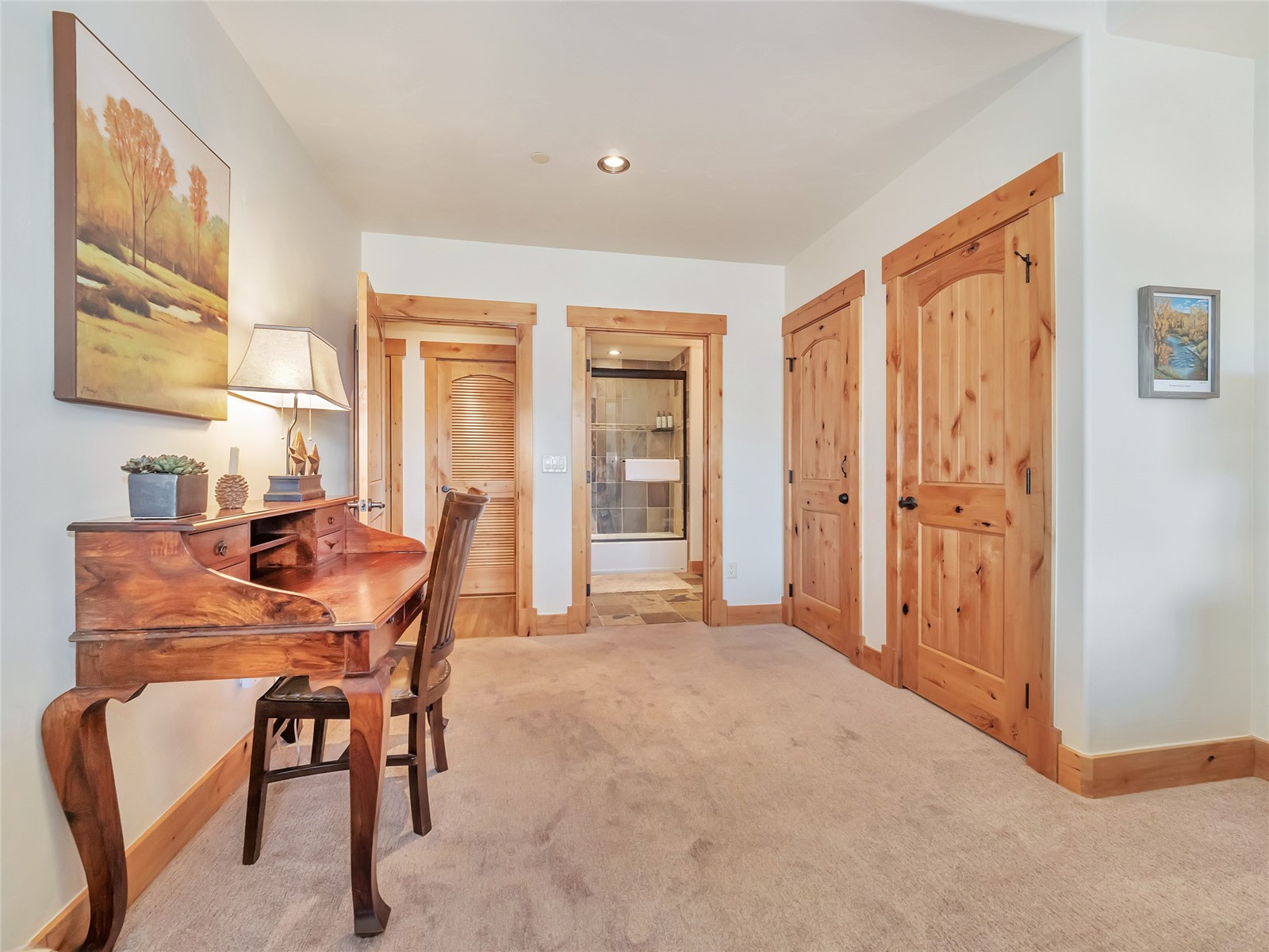 Carpeted hallway with a wooden writing desk, knotty wood doors, and an open view into a tiled bathroom.