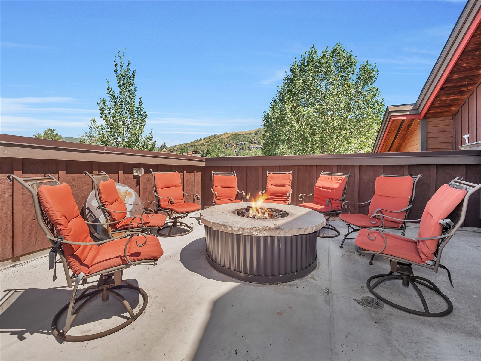 Patio with orange-cushioned chairs around a fire pit, overlooking a tree-lined hillside.