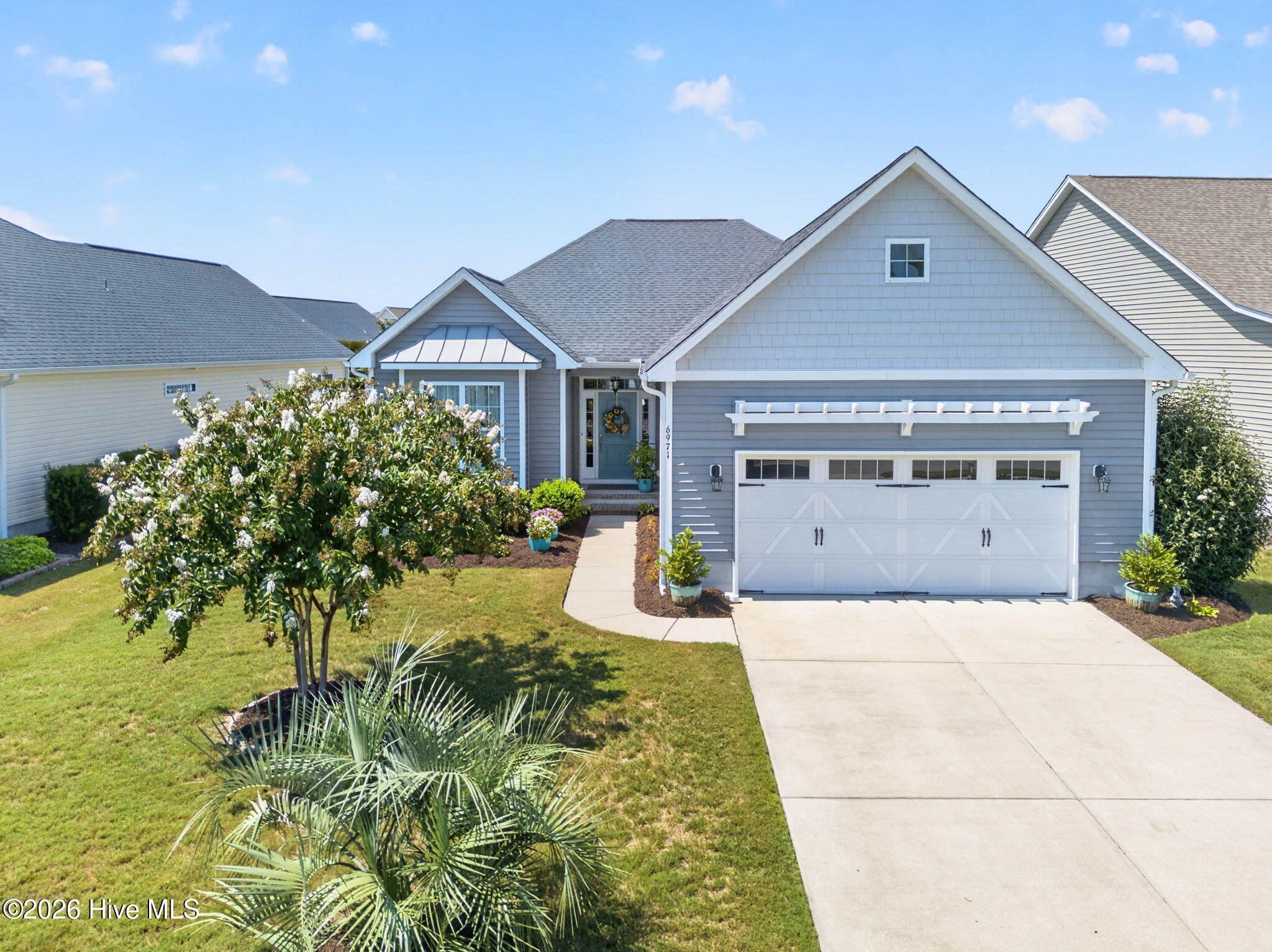 Gray shingle-style house with a white double garage, flowering shrubs, and a broad concrete driveway.