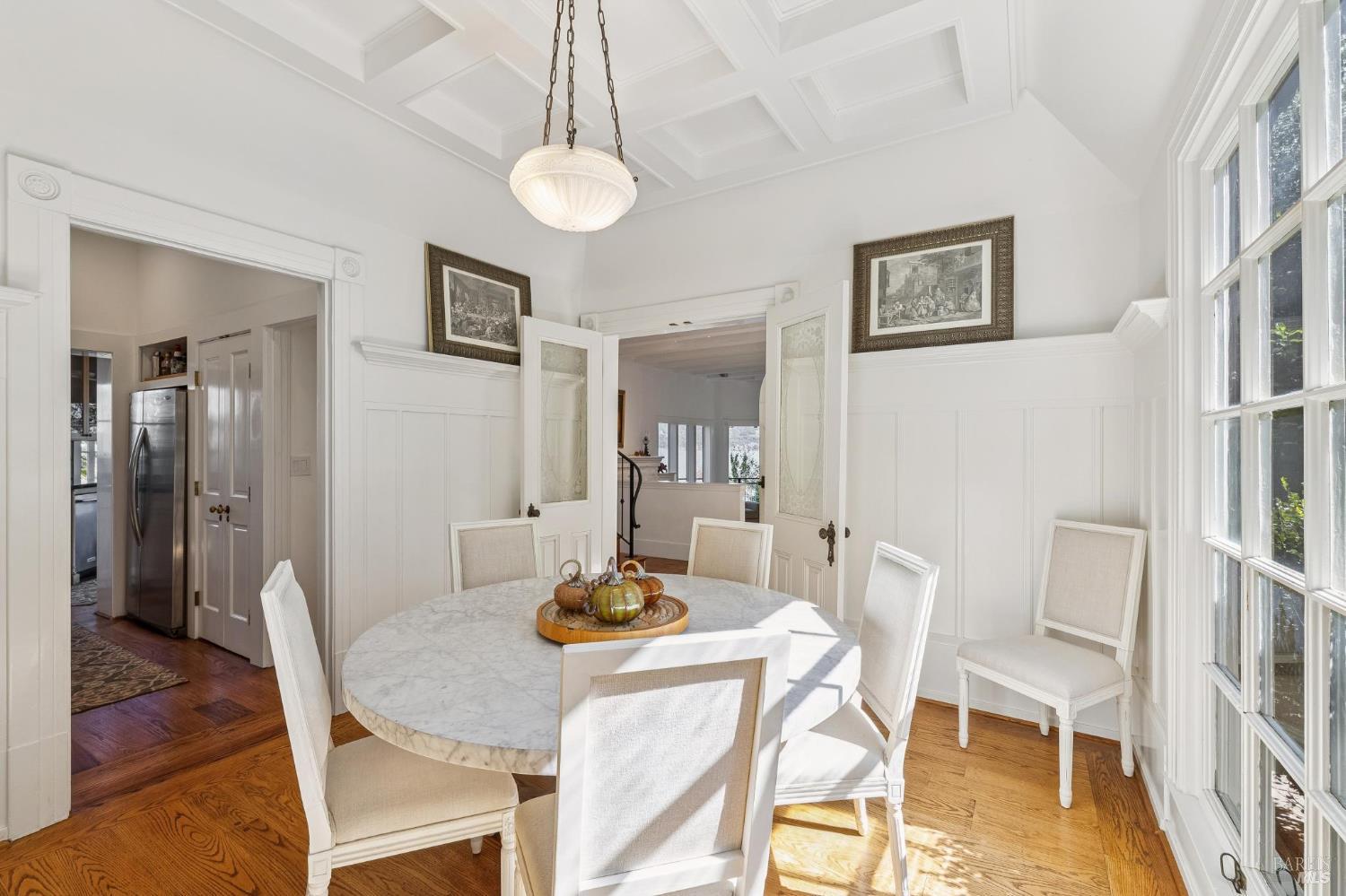Dining room with a marble-top table, white paneled walls, and tall sunlit windows.