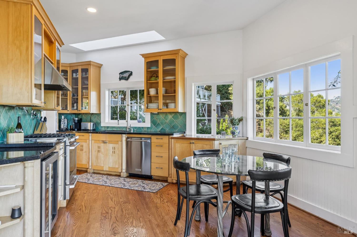 Bright kitchen with honey-colored cabinets, turquoise tile backsplash, and a glass dining table beside large windows.