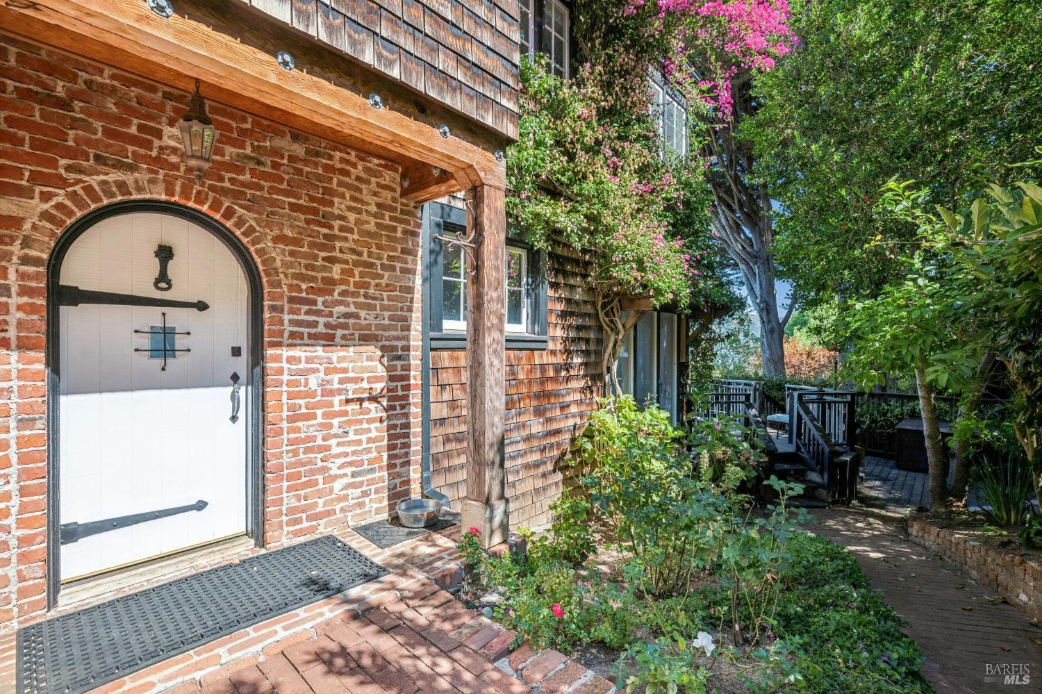 Brick-and-shingle home entrance with an arched white door, flowering vines, and a garden walkway.