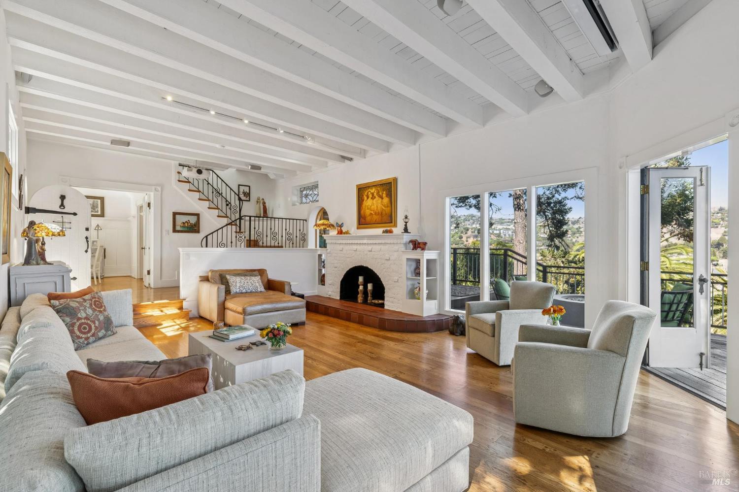 Living room with exposed ceiling beams, a white brick fireplace, and doors opening to a hillside view.