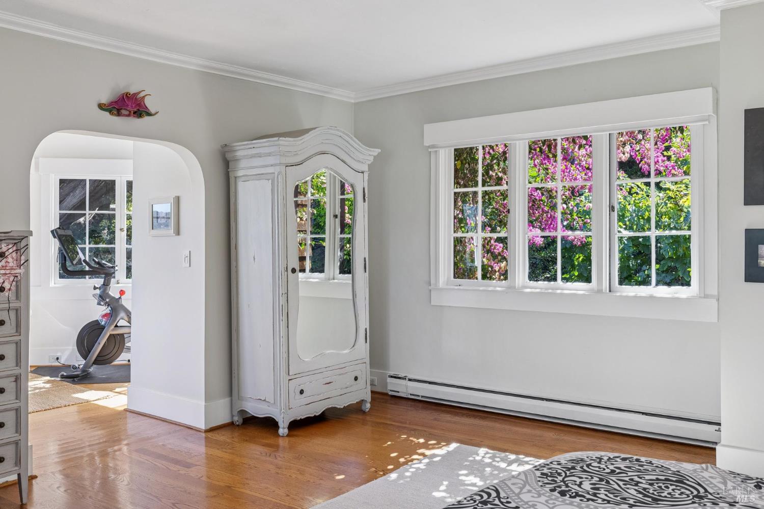 Bedroom with a white mirrored armoire, hardwood floors, and windows overlooking flowering shrubs.