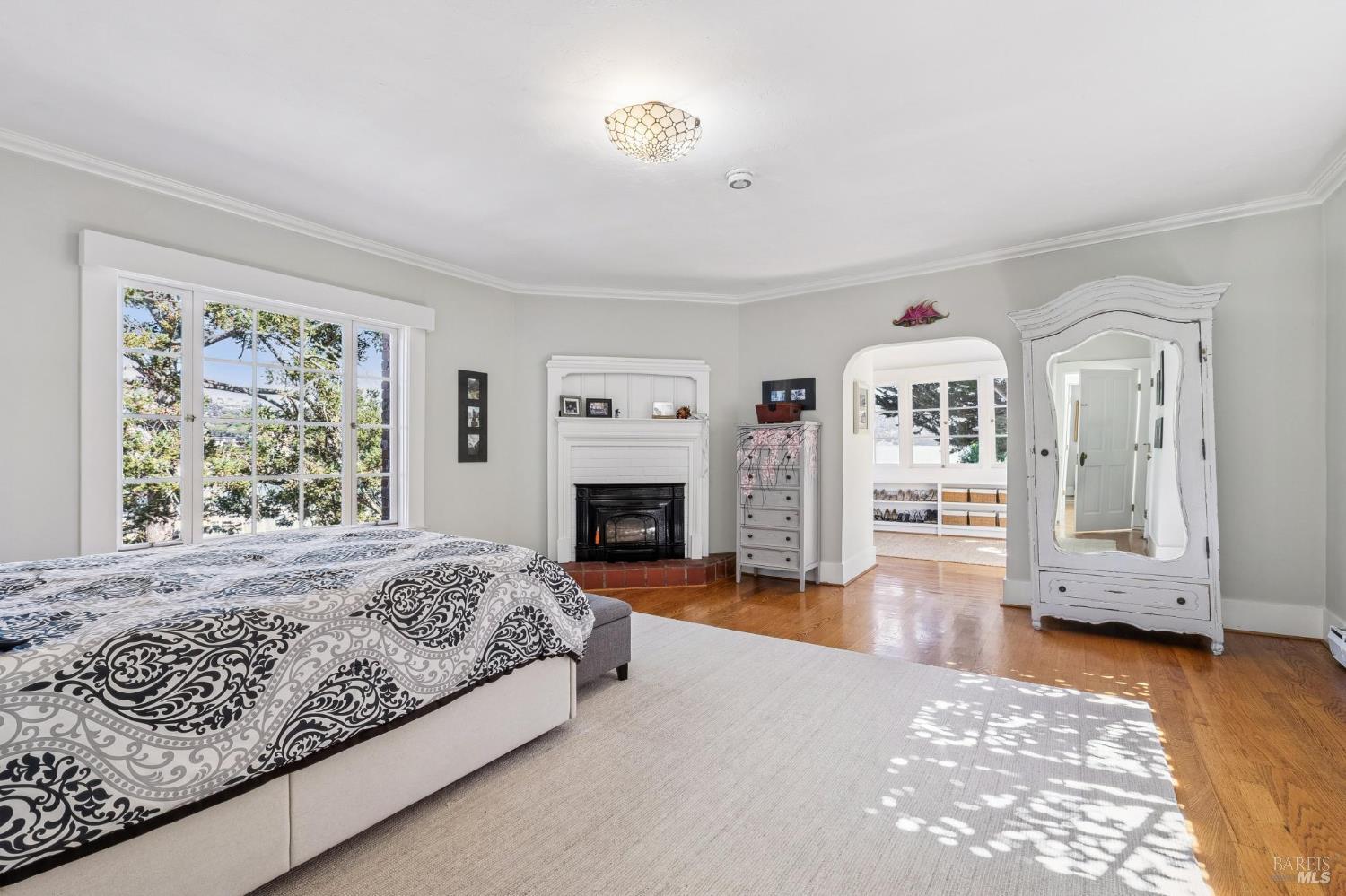 Bedroom with a patterned bedspread, white fireplace, hardwood floors, and large windows.