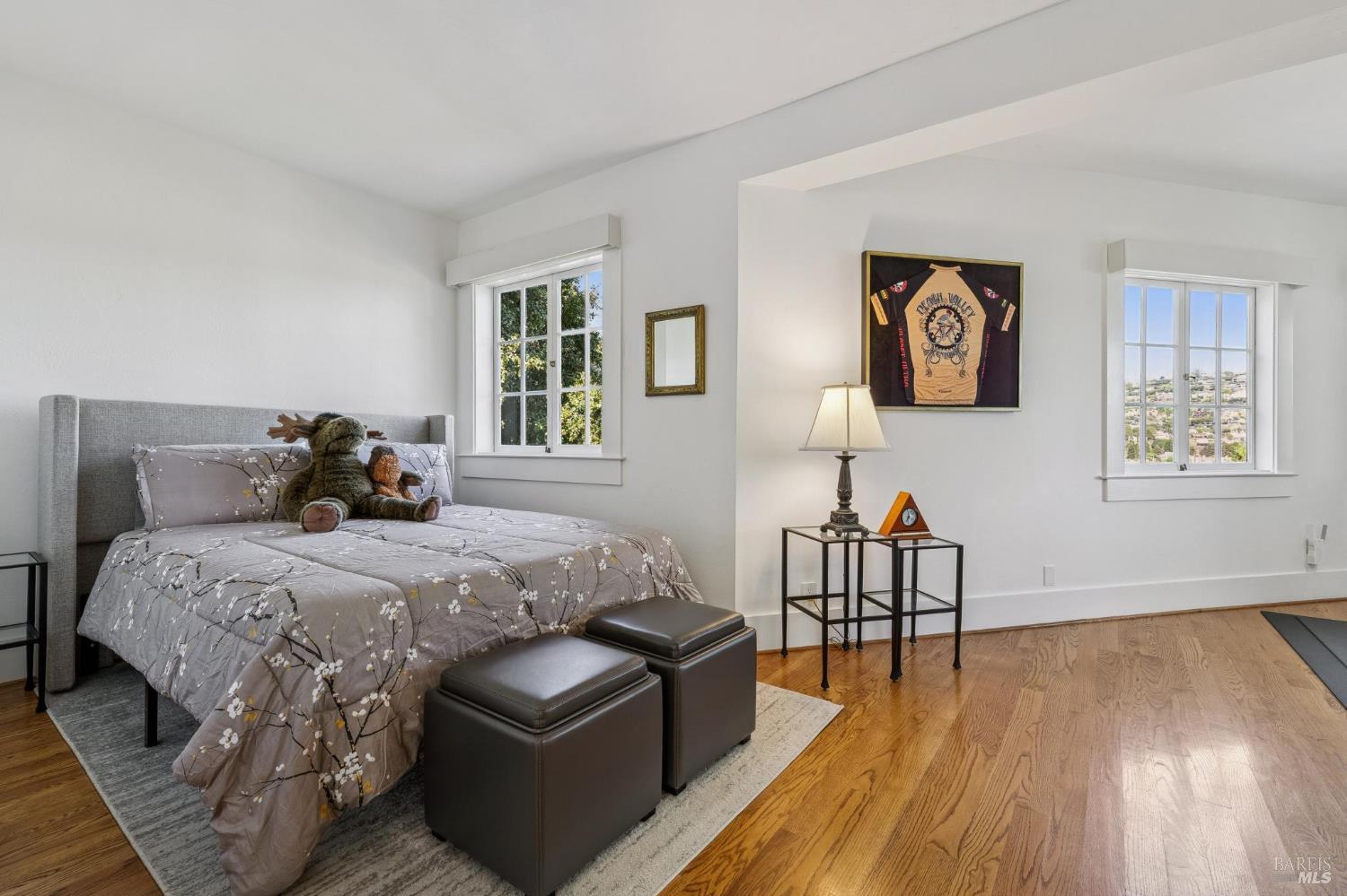 Bedroom with a gray upholstered bed, hardwood floors, and windows overlooking the neighborhood.