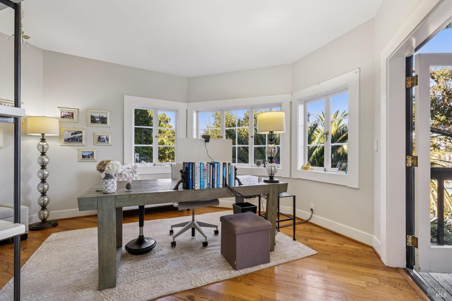 Bright home office with a desk, books, and windows overlooking palm trees.