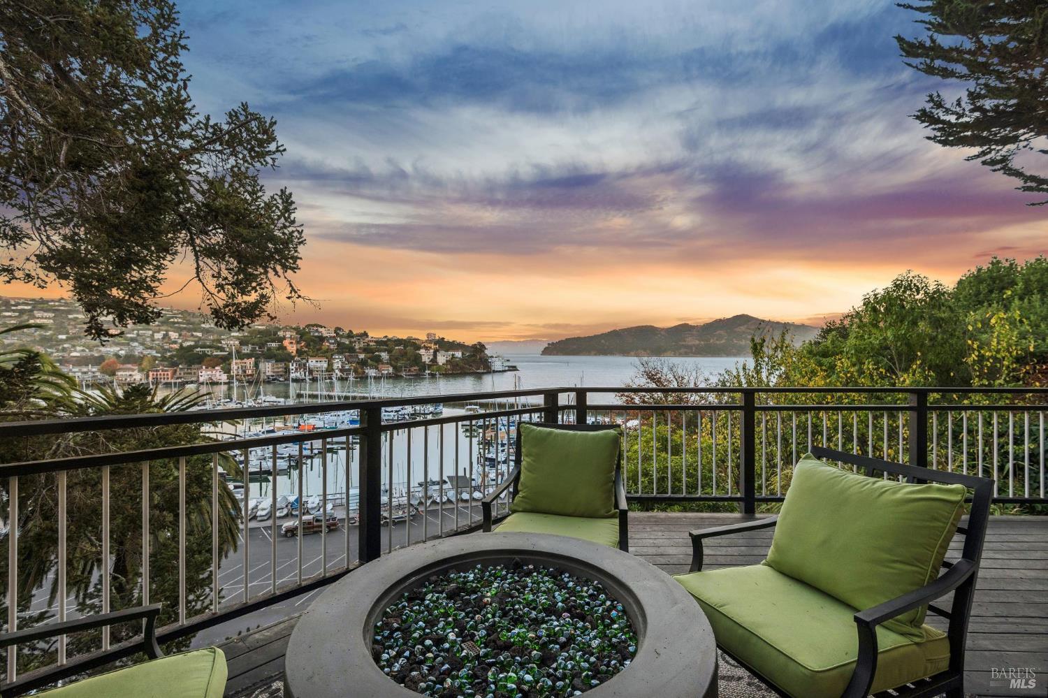 Furnished deck overlooking a marina and bay at sunset, with a circular fire pit and green lounge chairs.