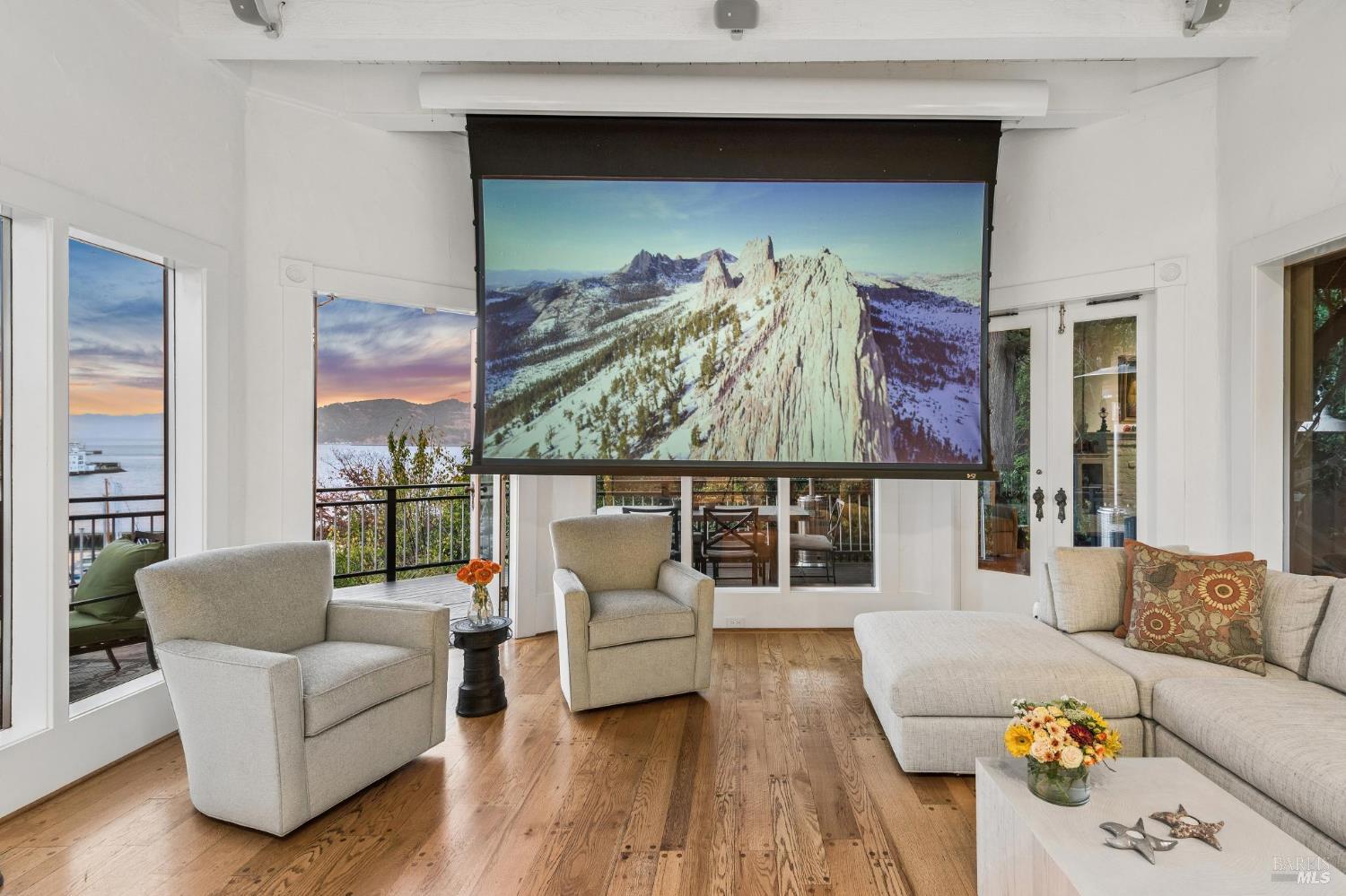 Living room with a large projection screen, light furnishings, and water views through the windows.