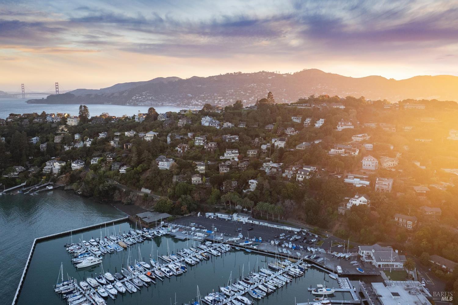Aerial view of a marina and hillside homes at sunset, with the Golden Gate Bridge in the distance.