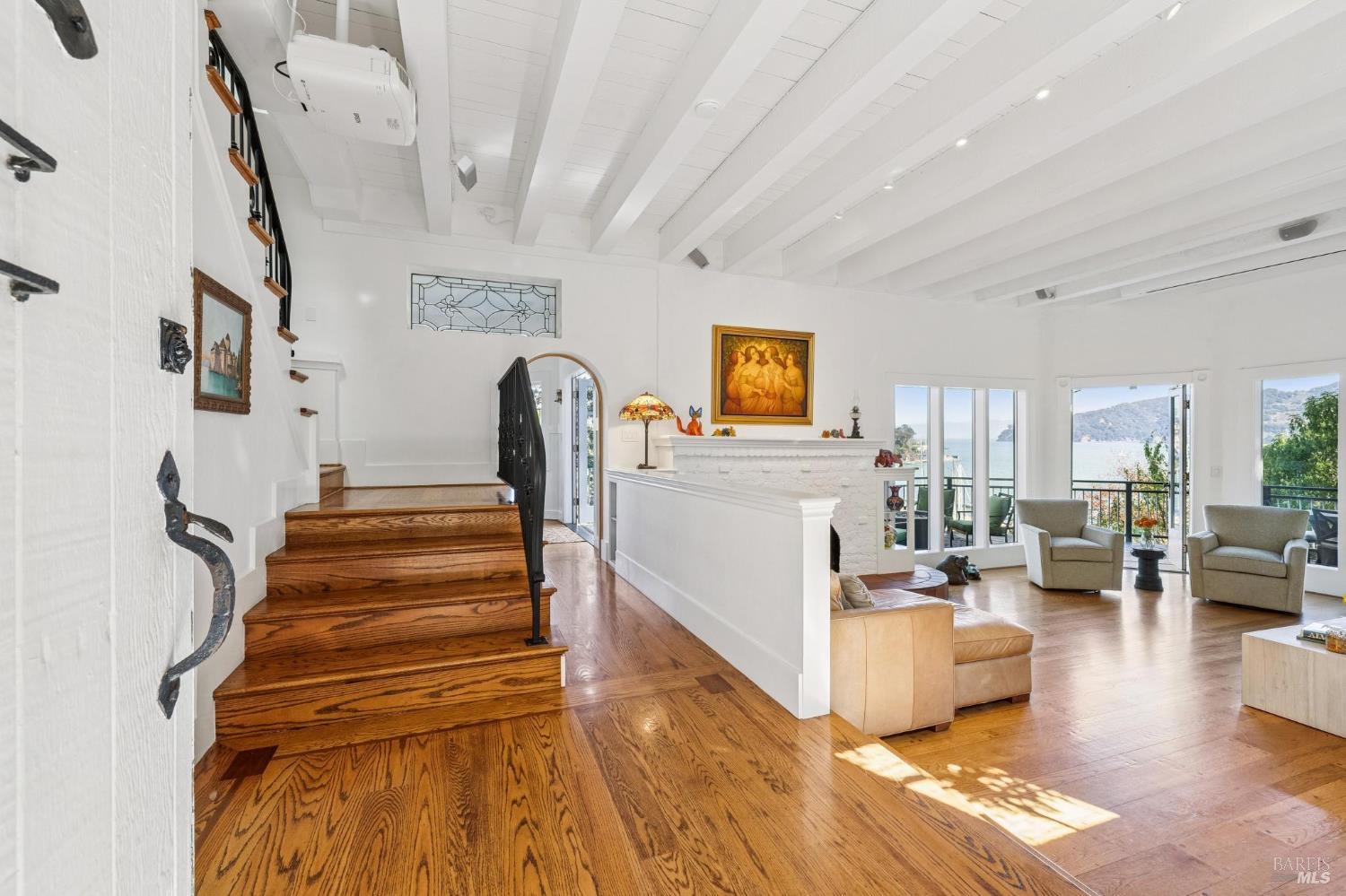 Living room with a wood staircase, exposed ceiling beams, and views of the water through open doors.