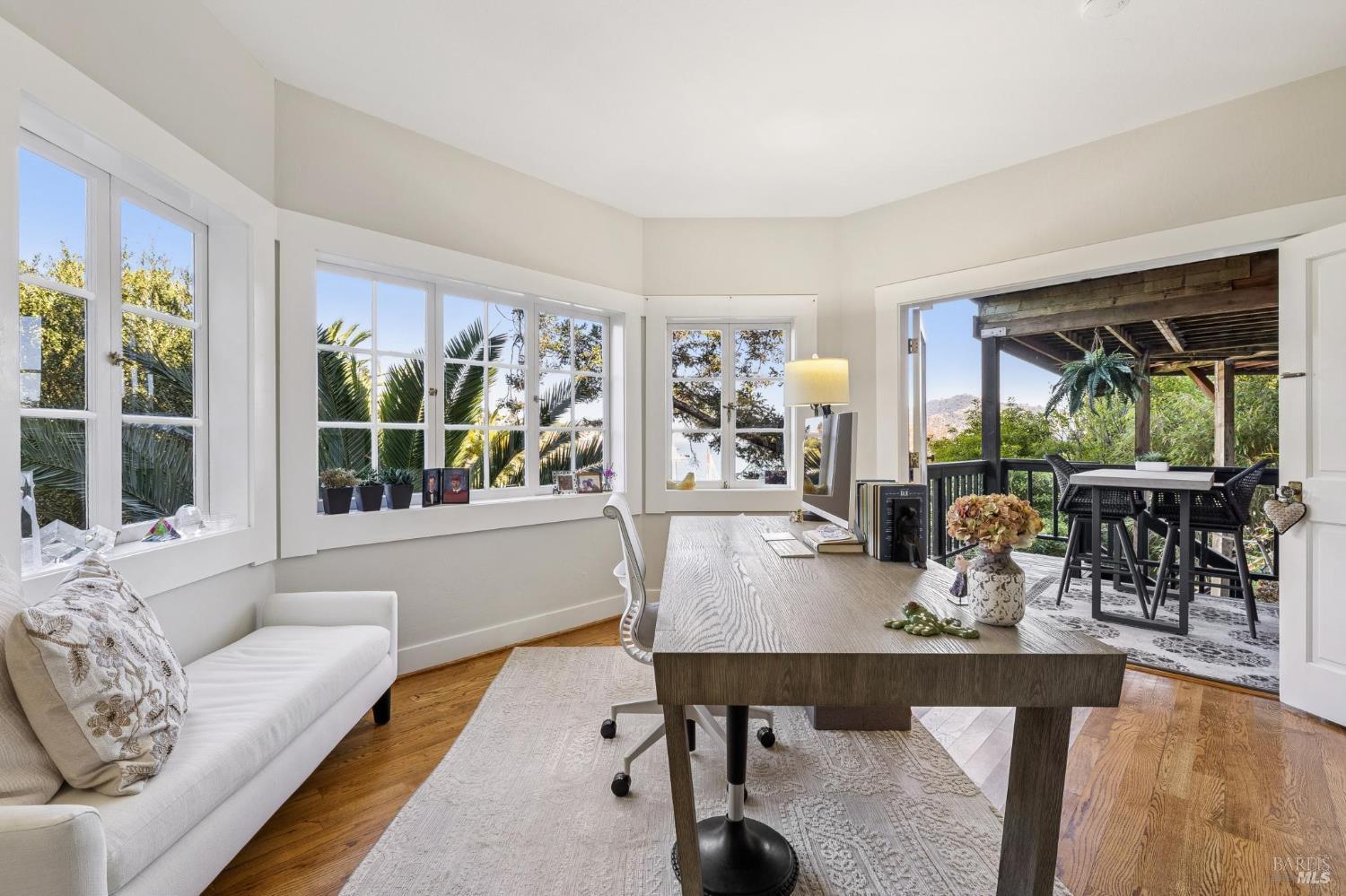 Furnished home office with a long desk, white sofa, and wide windows opening to a covered deck.