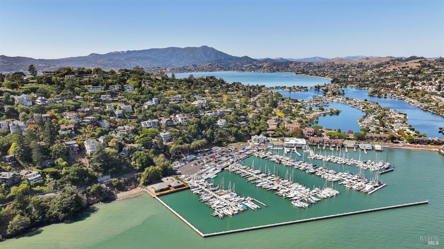 Aerial view of a marina with sailboats, shoreline homes, and hills across the bay.