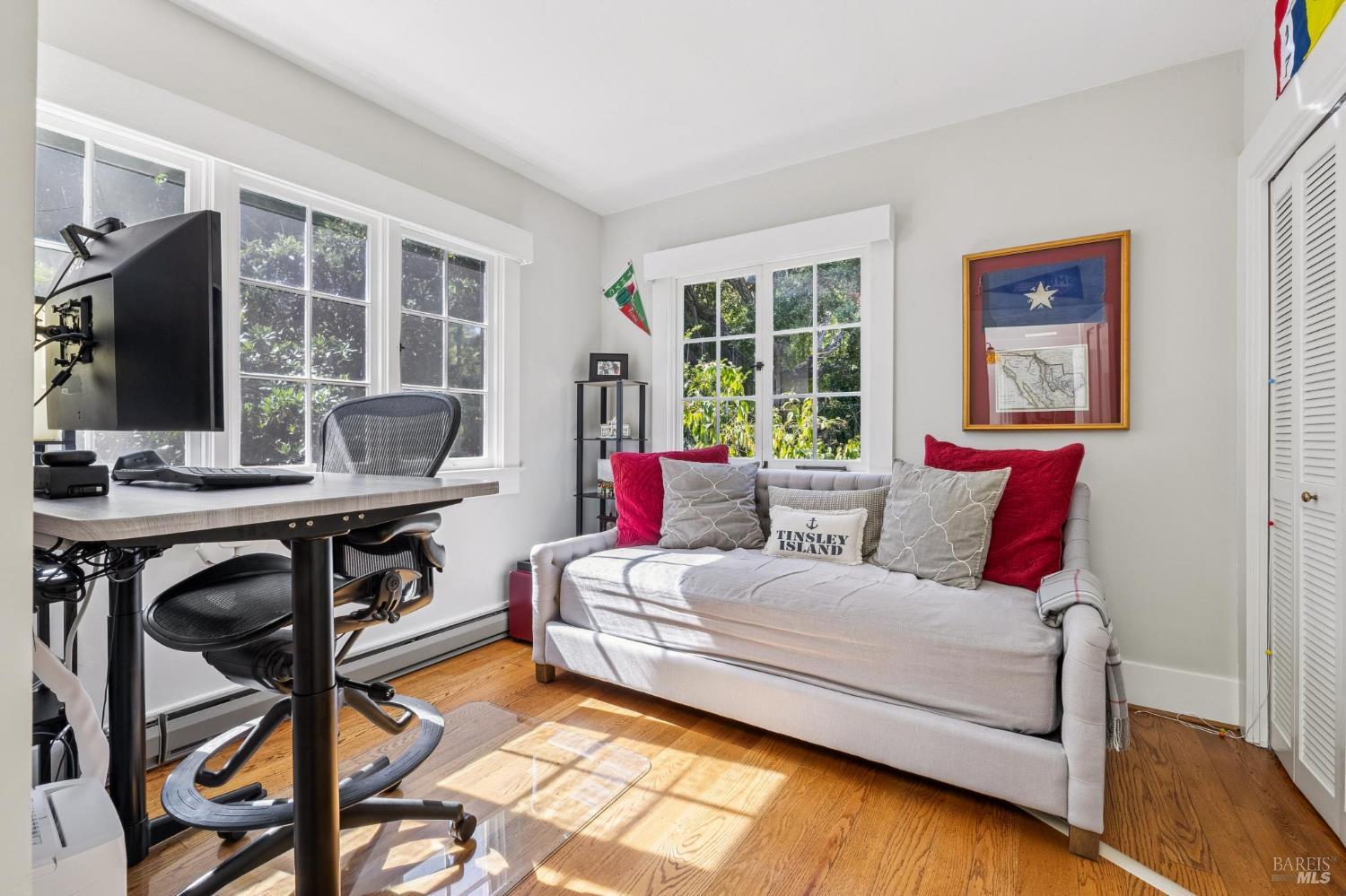 Home office with a desk beside a gray sofa and sunlit windows.
