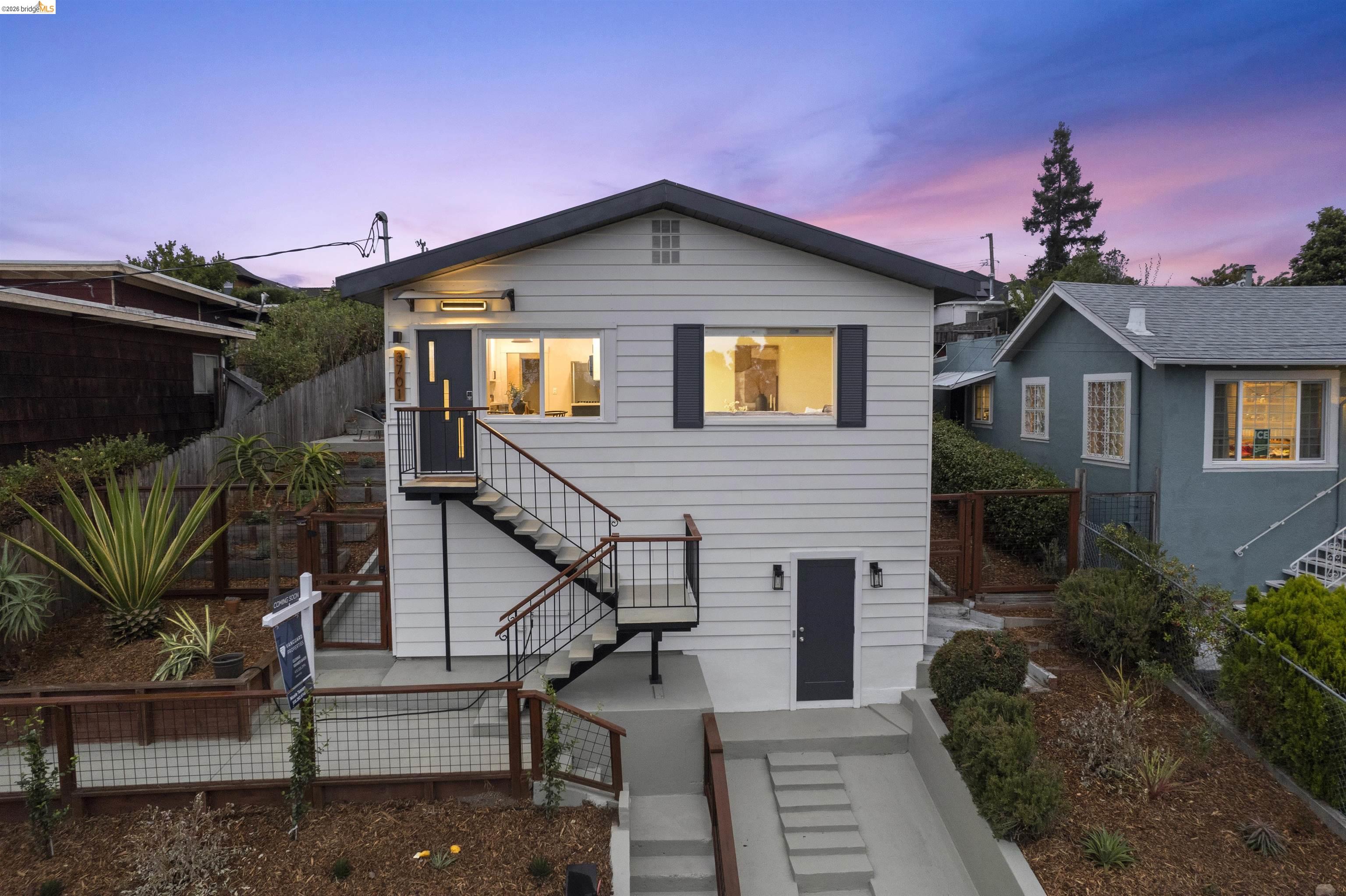 White Oakland home at dusk, with illuminated windows, exterior stairs, and a landscaped sloping yard.