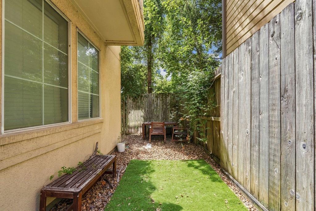 Fenced backyard with a strip of turf, wooden benches, and a small seating area beneath leafy trees.