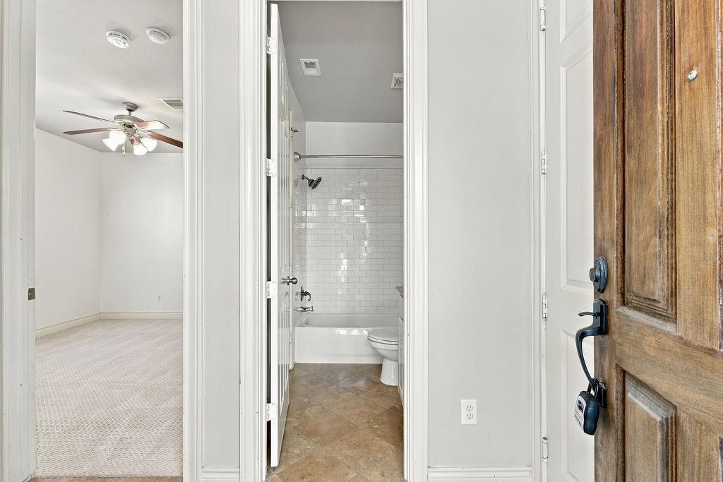 Entryway view of a tiled bathroom beside an unfurnished carpeted bedroom and rustic wood door.