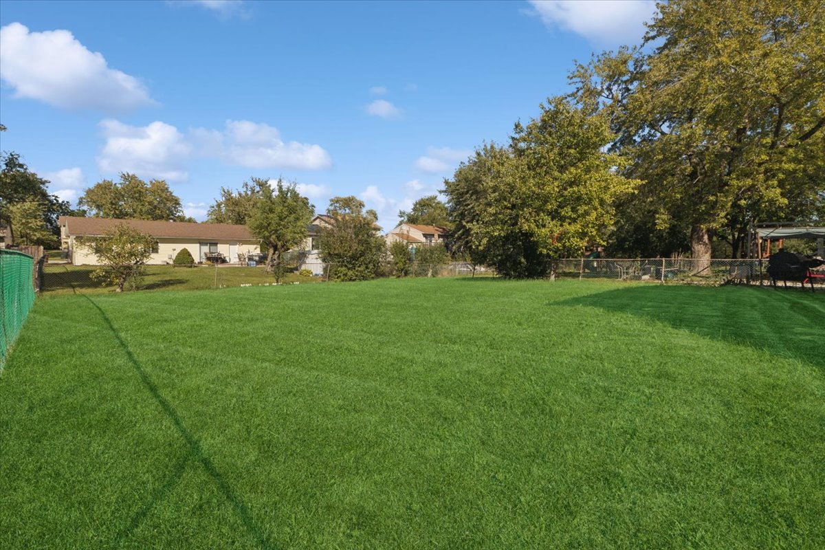 Broad grassy backyard with mature trees, fencing, and neighboring homes in the distance.