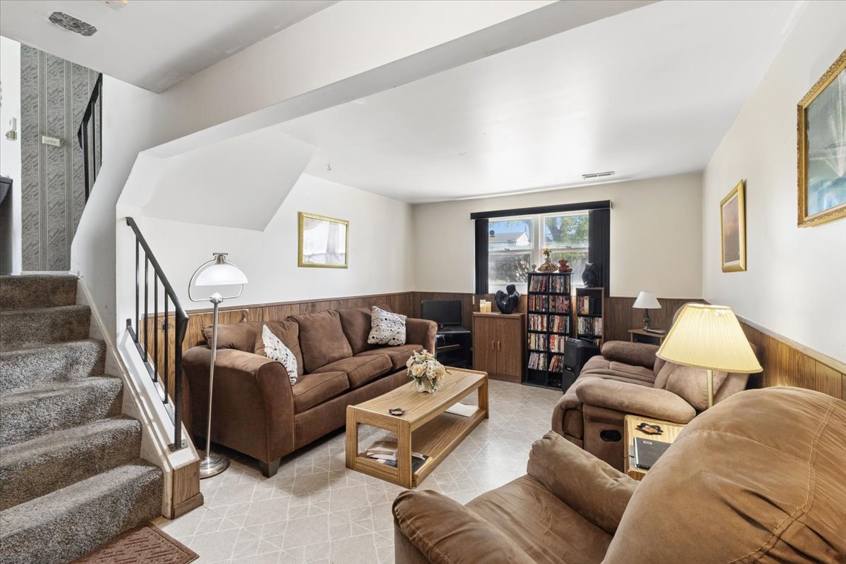 Living room with brown seating, wood-paneled walls, and a staircase beside the seating area.