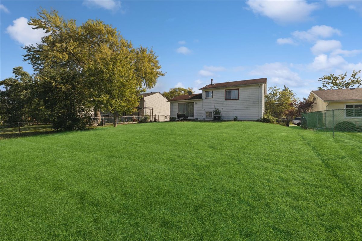 Rear of a two-story house beyond a broad lawn, with mature trees and neighboring homes.
