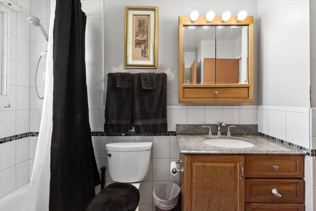 Bathroom with a black shower curtain, tiled walls, and a wood vanity with granite counter.