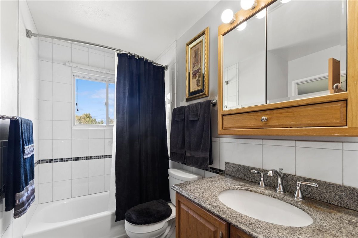 Bathroom with a white tub, dark shower curtain, and wood vanity topped with a granite counter.