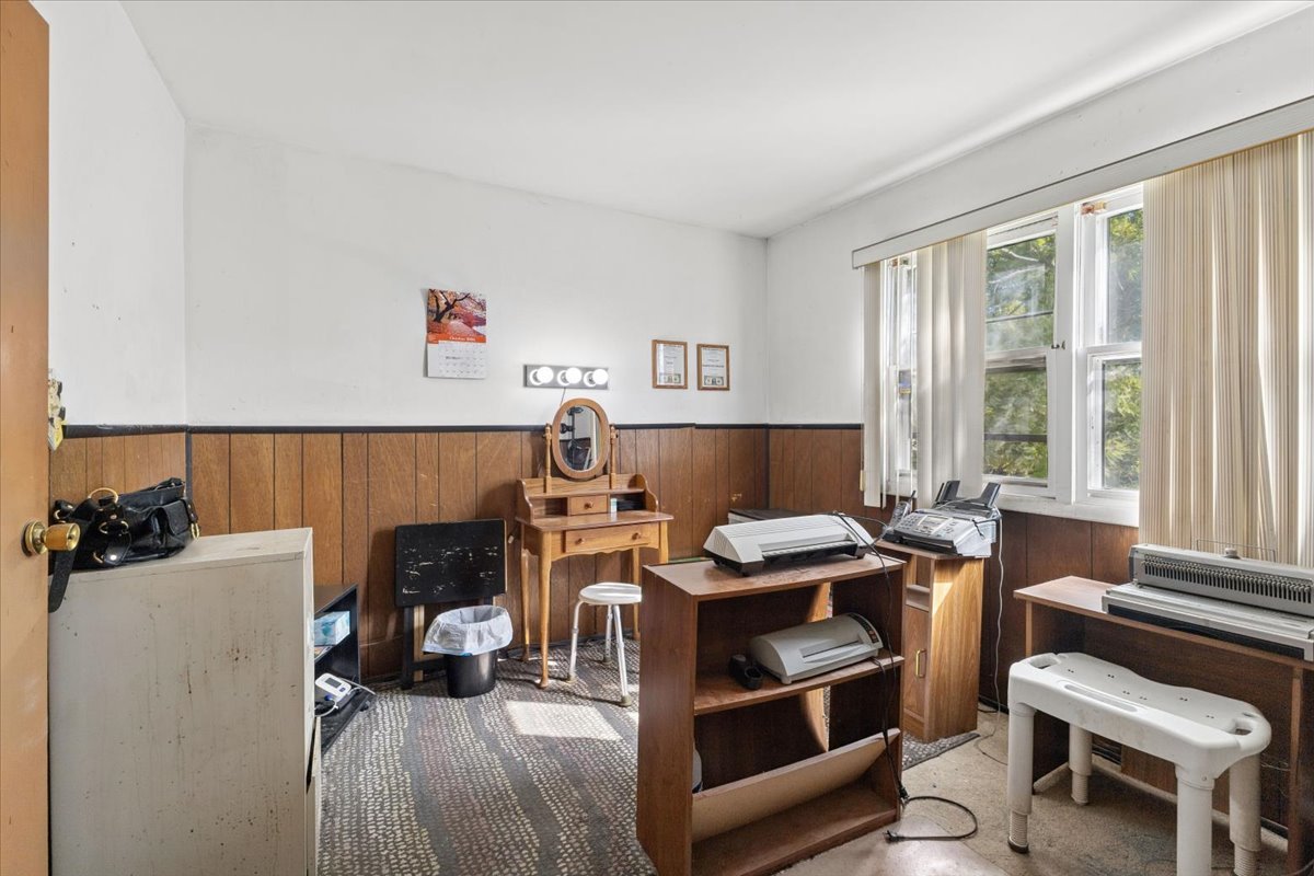 Cluttered room with wood paneling, desks, equipment, and tall windows with vertical blinds.