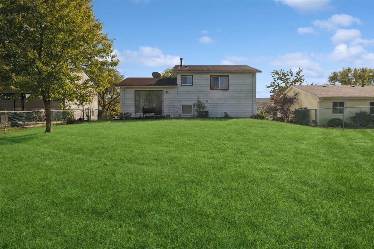 Rear of a two-story house beyond a broad lawn, with chain-link fencing and neighboring homes.
