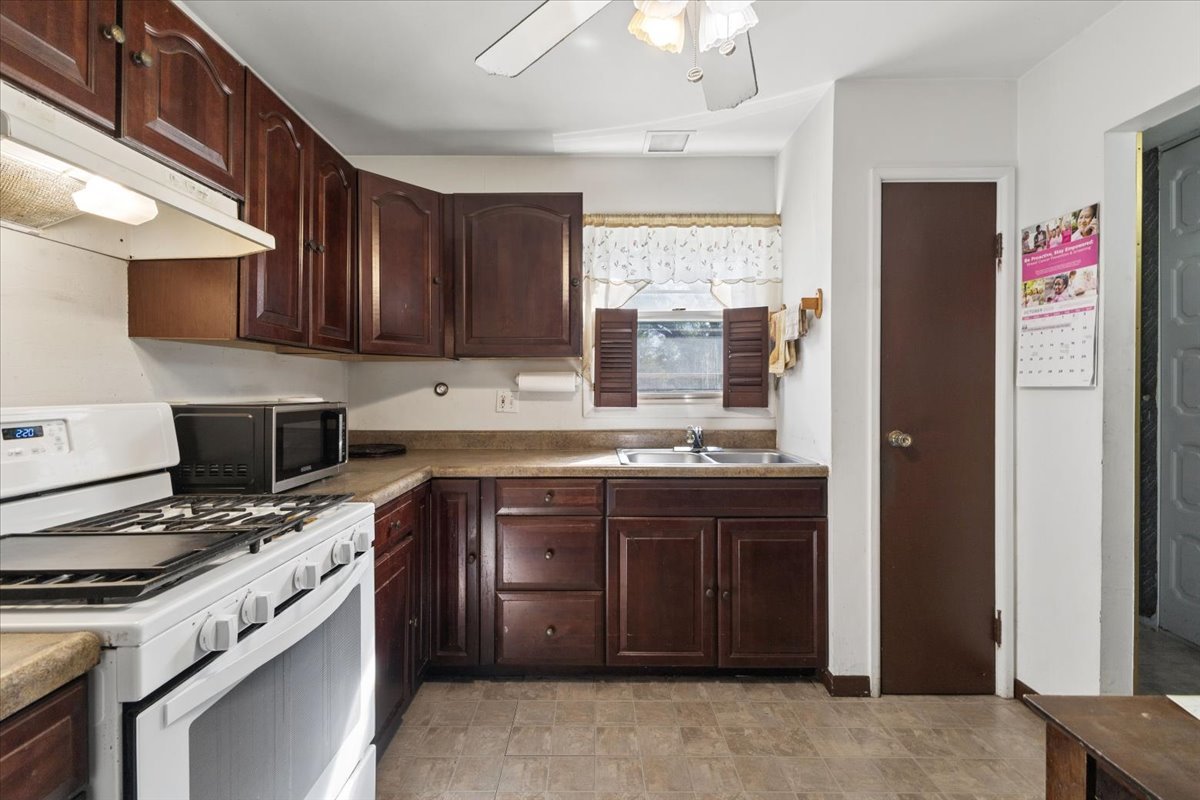 Kitchen with dark wood cabinets, tan counters, white gas range, and a sink beneath a shuttered window.