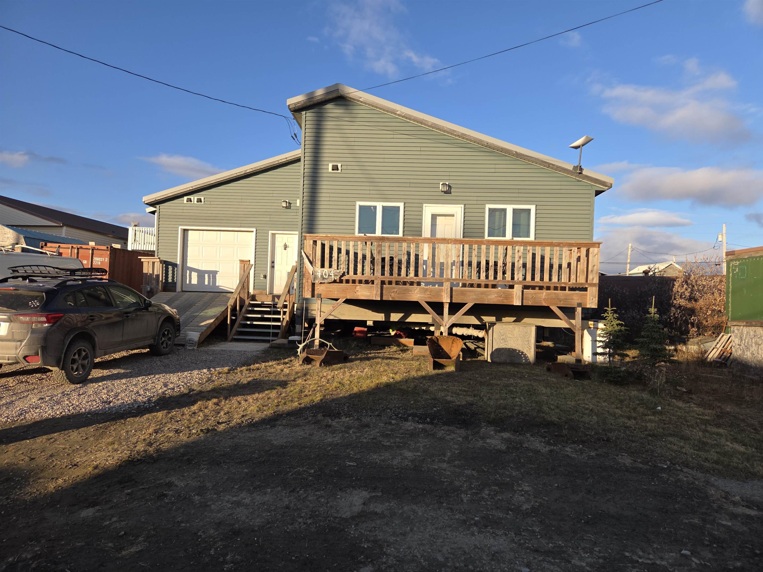 Gray-green house with an elevated wooden deck, exterior stairs, and a gravel driveway.