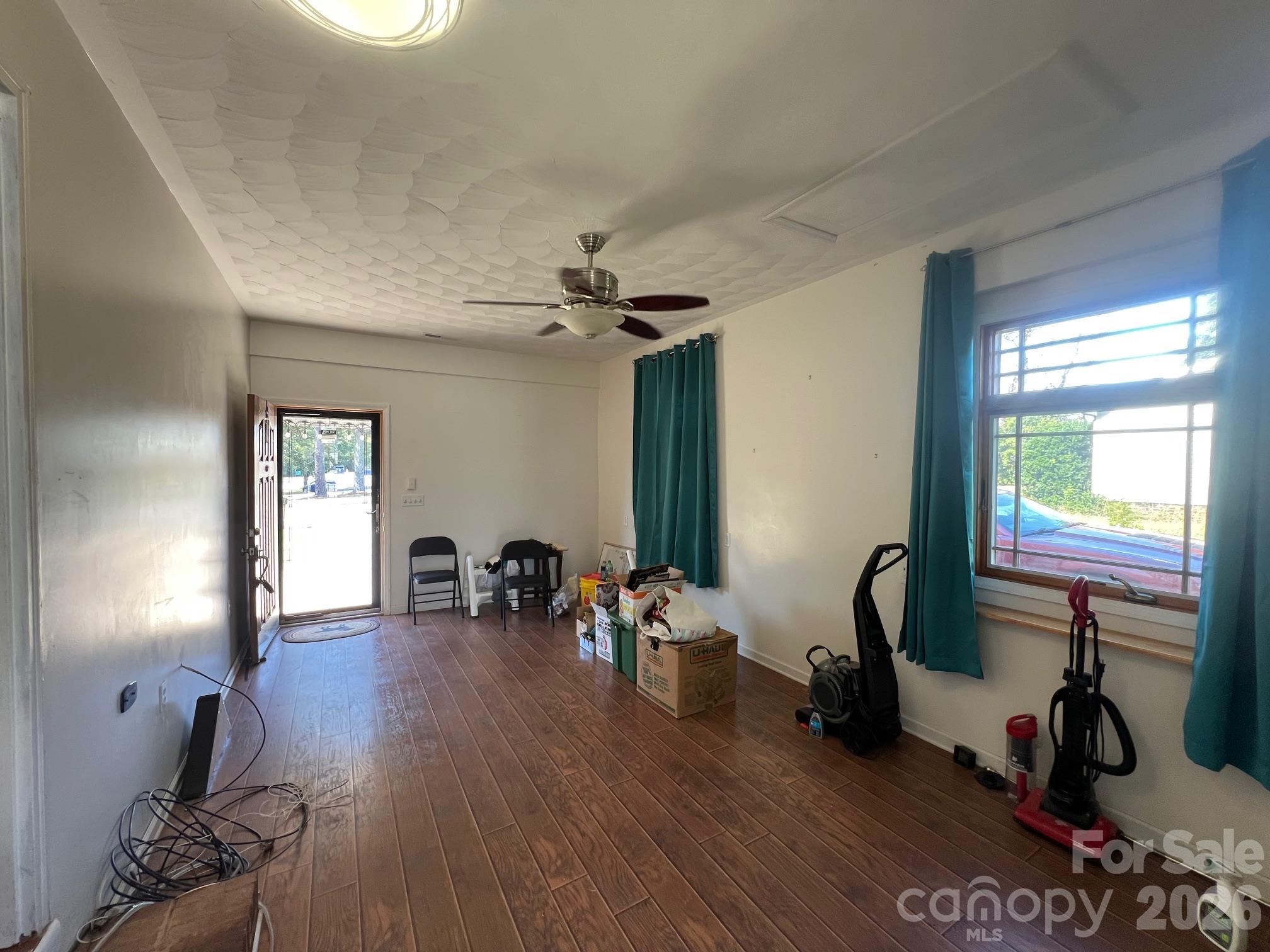 Long room with brown plank flooring, teal curtains, scattered boxes, chairs, and cleaning equipment.