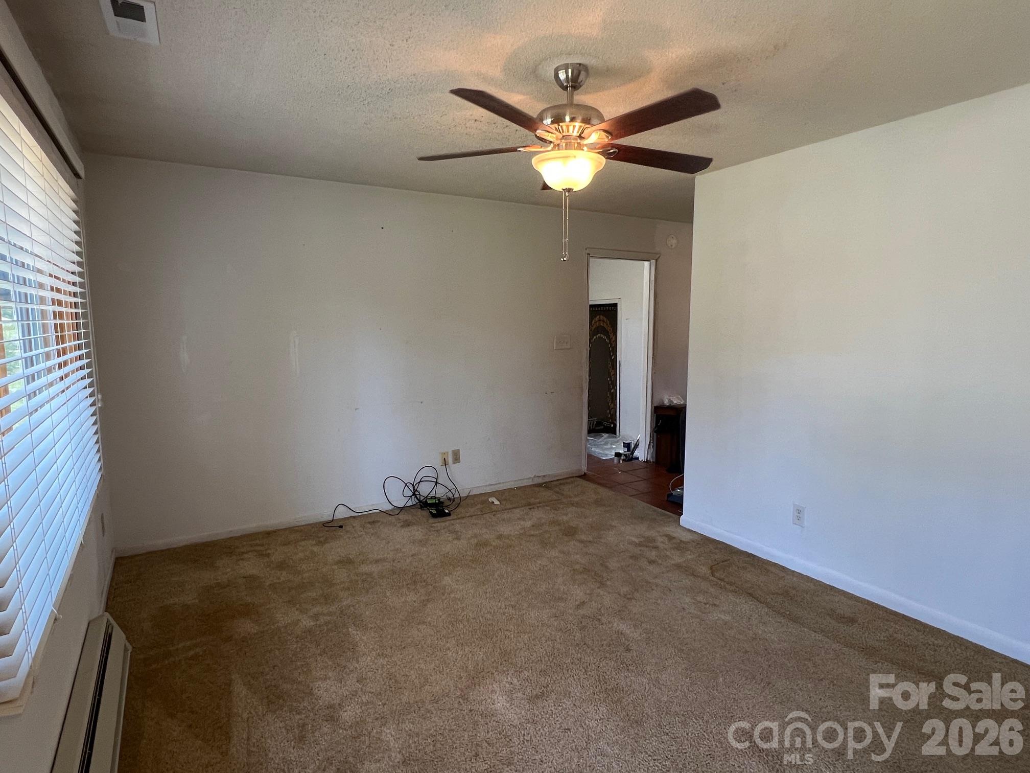 Empty carpeted living room with a lit ceiling fan, wide window with blinds, and doorway to a tiled area.